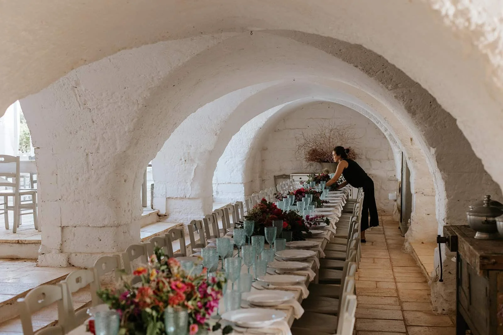 Woman arranging floral centerpieces along long dinner table inside barrel-vault stone cellar at Masseria Potenti