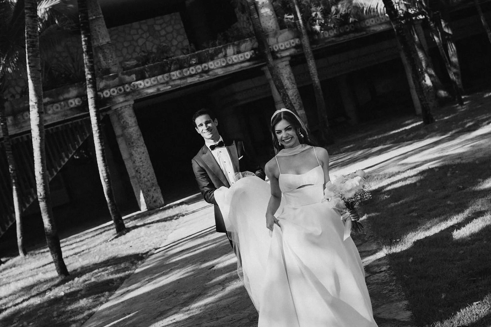 Bride and groom smiling during couple portraits at Xcaret Park in Playa Del Carmen, Mexico — black and white wedding photography with Mayan architecture and palm trees in background