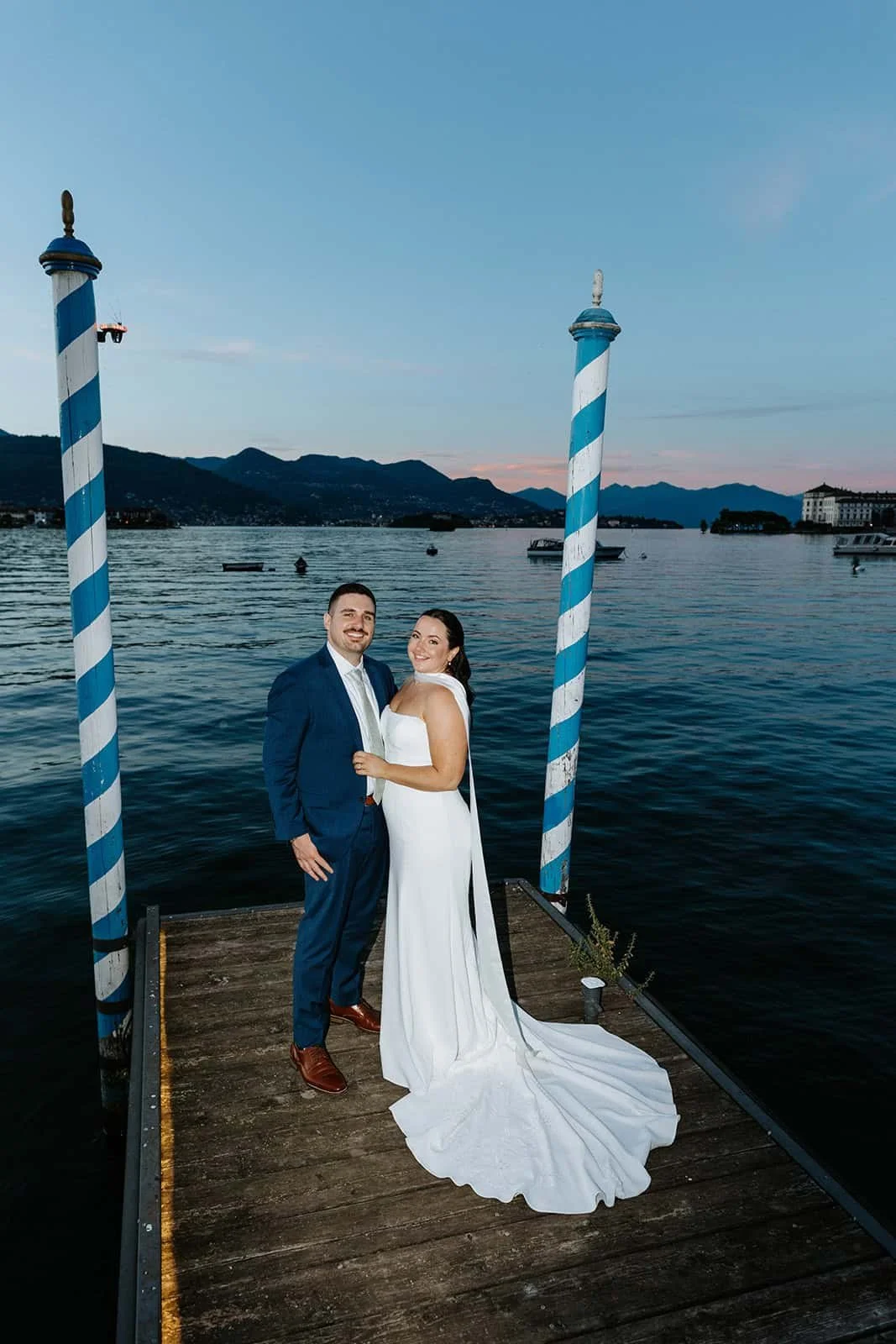 Erin and Bill posing on the dock between blue and white striped mooring poles at Villa Aminta, with Lago Maggiore, mountains and town of Stresa in the background.