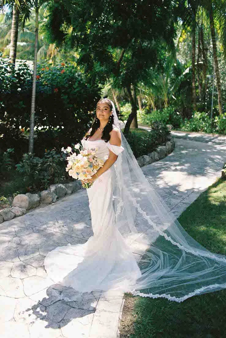 Bride in a white wedding gown with a long veil, holding a bouquet, standing on a paved pathway in a lush, green garden.
