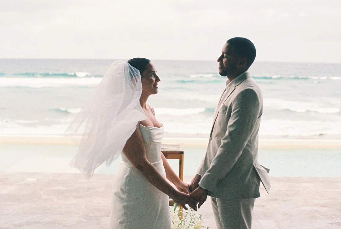 A bride and groom holding hands during their wedding ceremony on the beach with the ocean in the background.