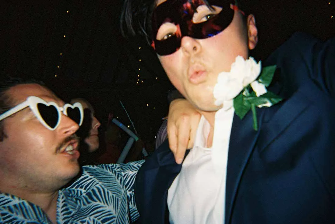 Two men at a party wearing sunglasses, one with heart-shaped glasses and the other with a masquerade mask, in a dark setting with string lights, dressed in flashy outfits, with one wearing a flower and bowtie.