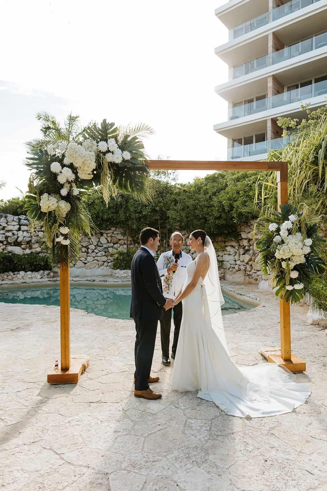 Couple at a wooden floral arch during their ceremony at the Mystique cenote area at Secrets Moxché Playa del Carmen, stone wall and tropical planting behind them