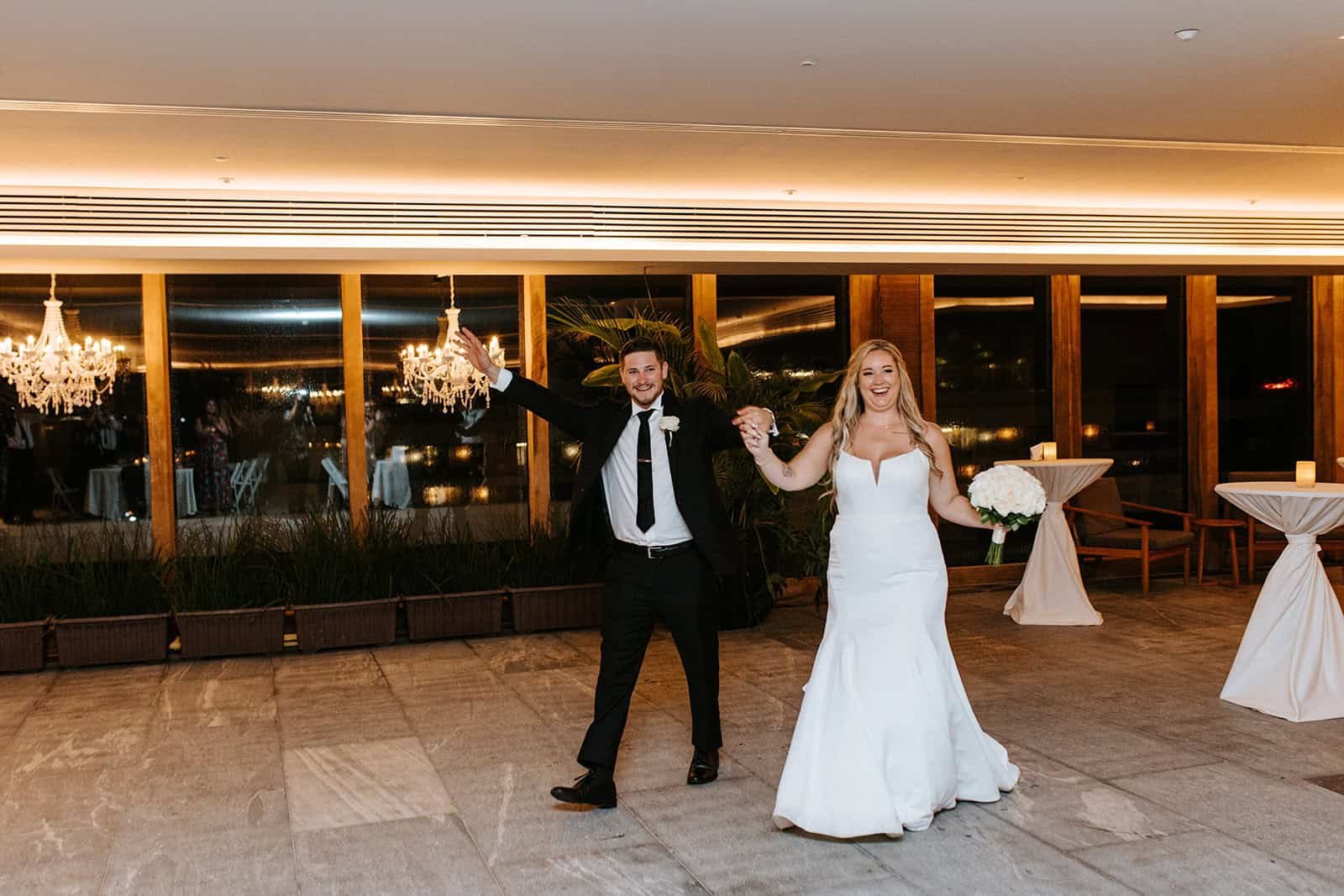 Couple entering their reception in the Sky Wedding Salón at Secrets Moxché Playa del Carmen, chandeliers and floor-to-ceiling windows behind them at night
