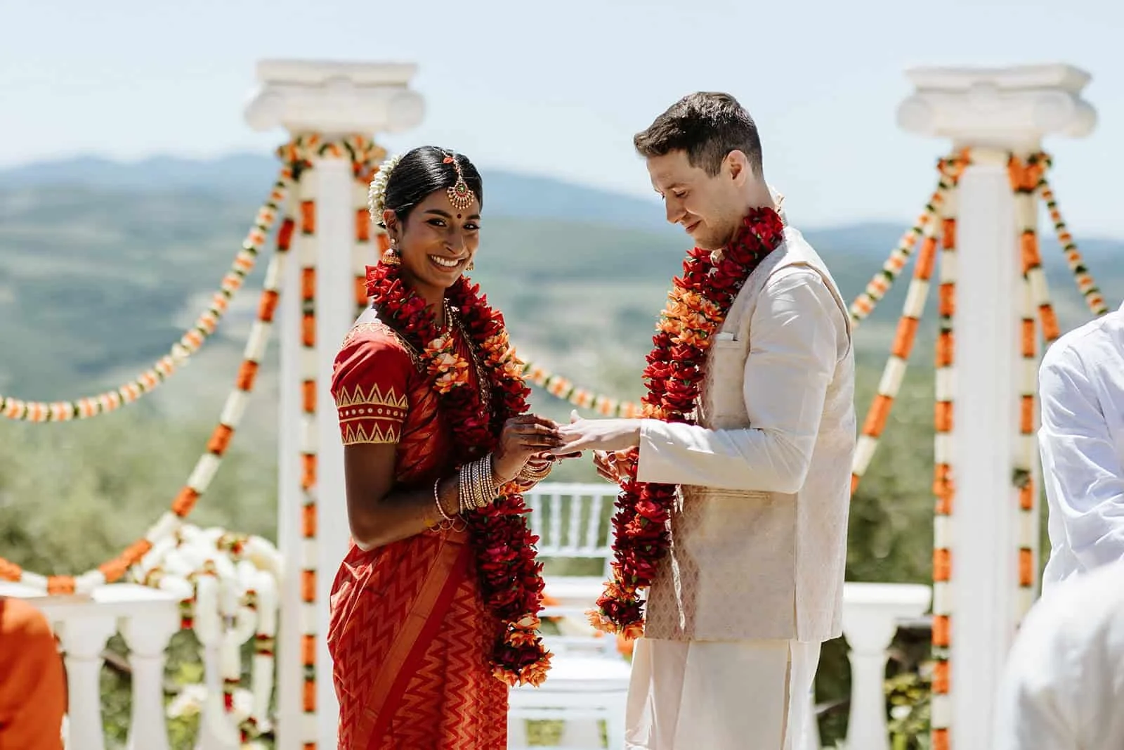 Hindu wedding ceremony at Todi, Umbria — bride in red saree and groom in white exchanging rings under marigold-decorated arch with valley views behind.