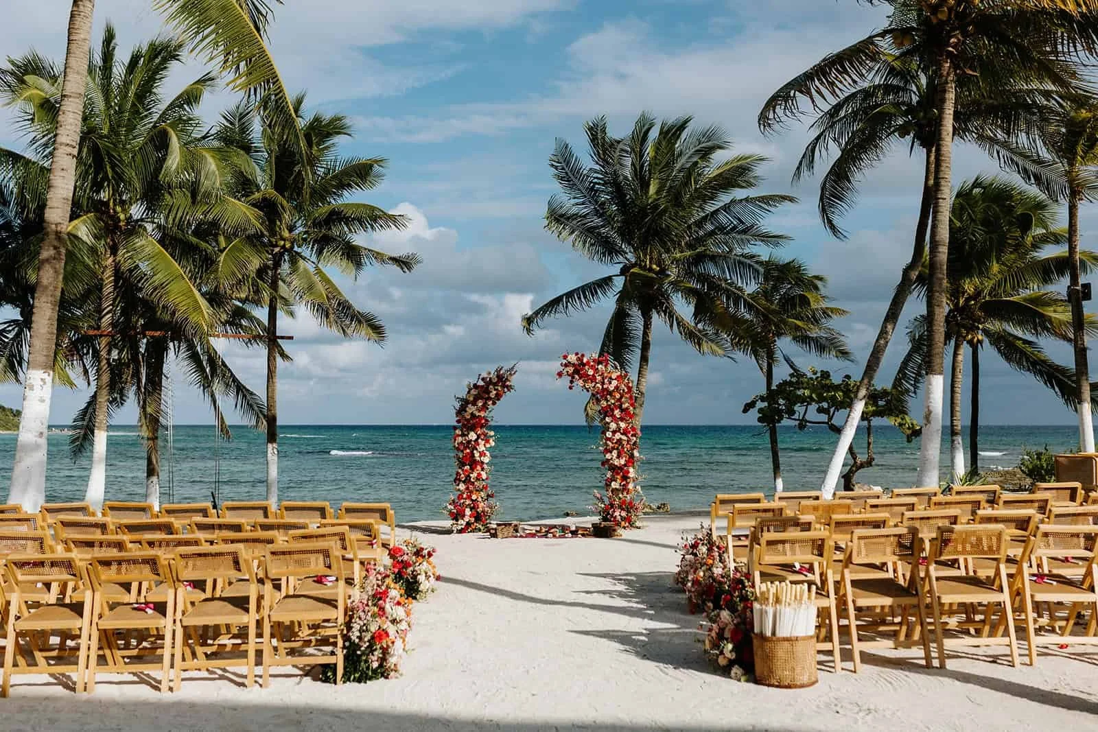 Beach wedding ceremony setup at Blue Venado Riviera Maya — open floral arch in red, blush and cream roses framing the Caribbean sea, rattan chairs lining the sandy aisle under palm trees