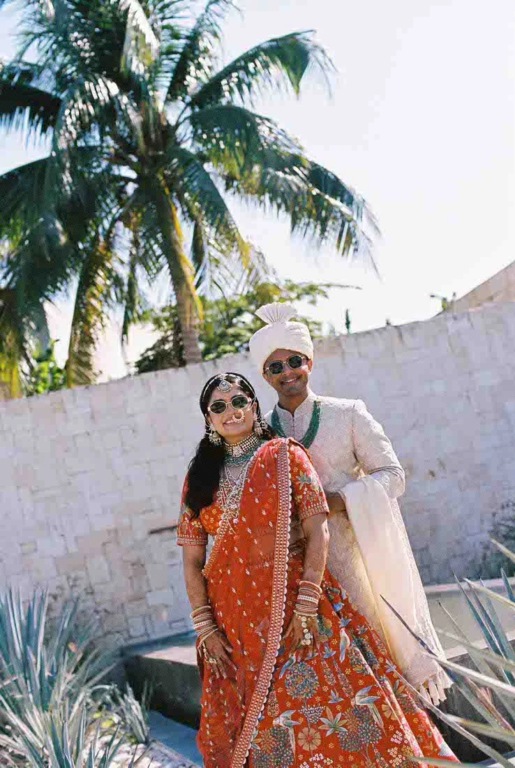 A couple dressed in traditional Indian wedding attire standing outdoors, with a white brick wall and palm trees in the background.