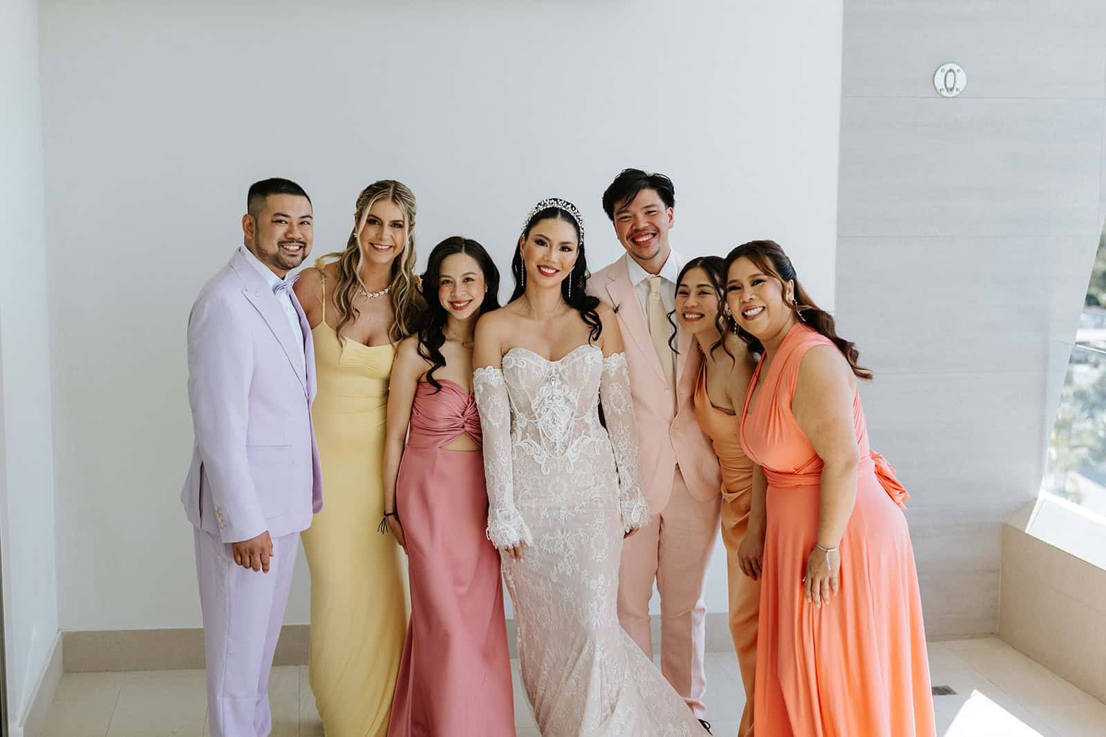 Bride Cathy in white lace dress with wedding party of bridesmaids and groomsmen in coordinated pastel and warm-toned outfits at Garza Blanca Cancun, all smiling at camera in modern white interior space