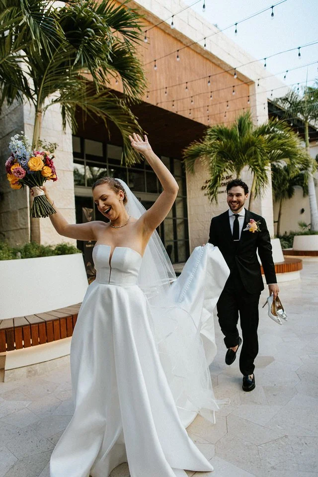 Bride and groom laughing as they exit their ceremony at Atelier Playa Mujeres, Cancún — groom carrying her heels