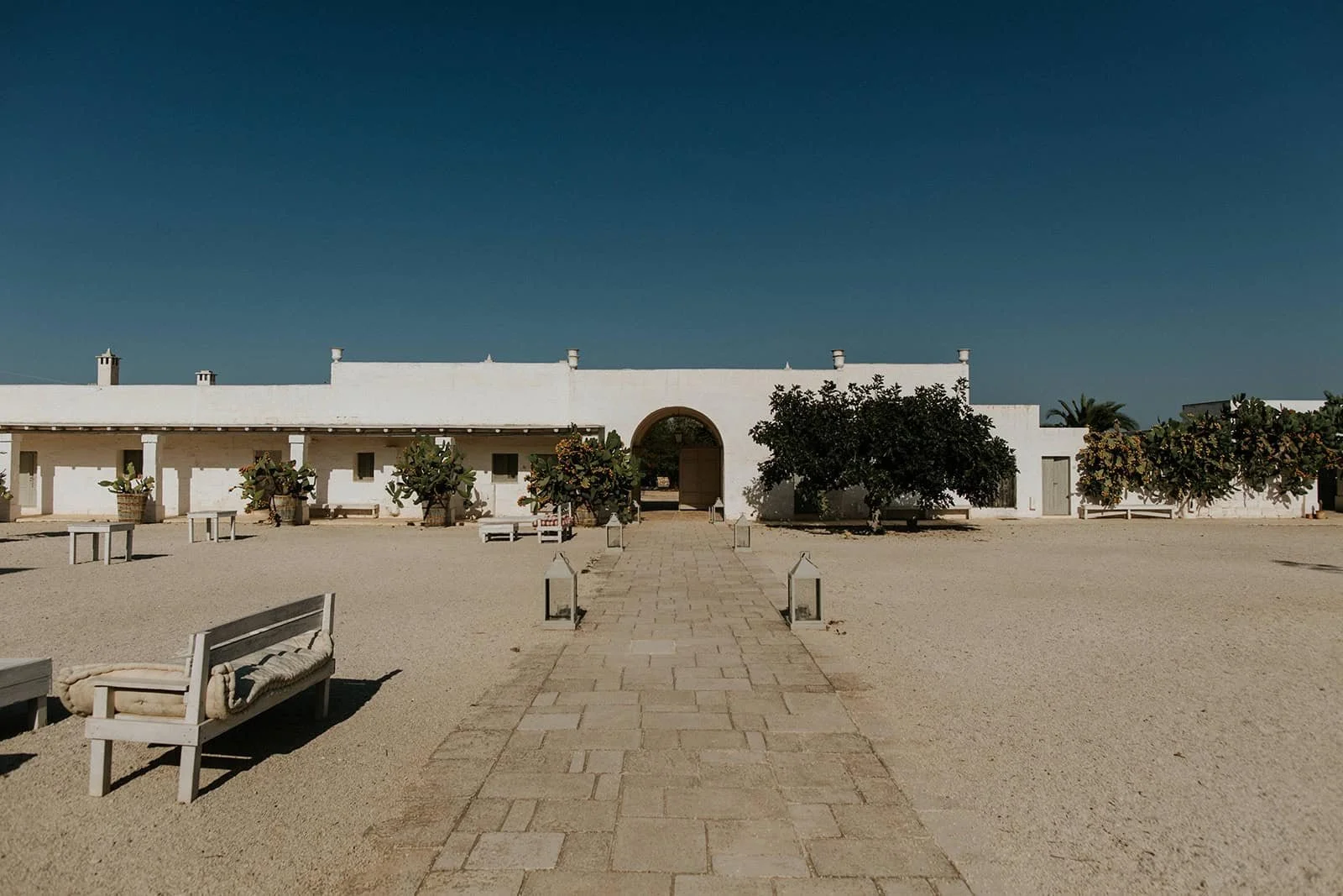 Wide view of Masseria Potenti main courtyard with stone pathway, whitewashed buildings, prickly pear cactus, and clear blue sky