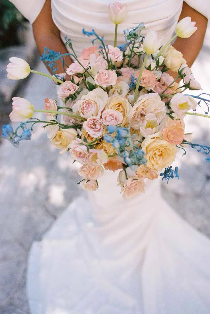 A woman in a white dress holding a large bouquet of light pink, cream, and blue flowers.