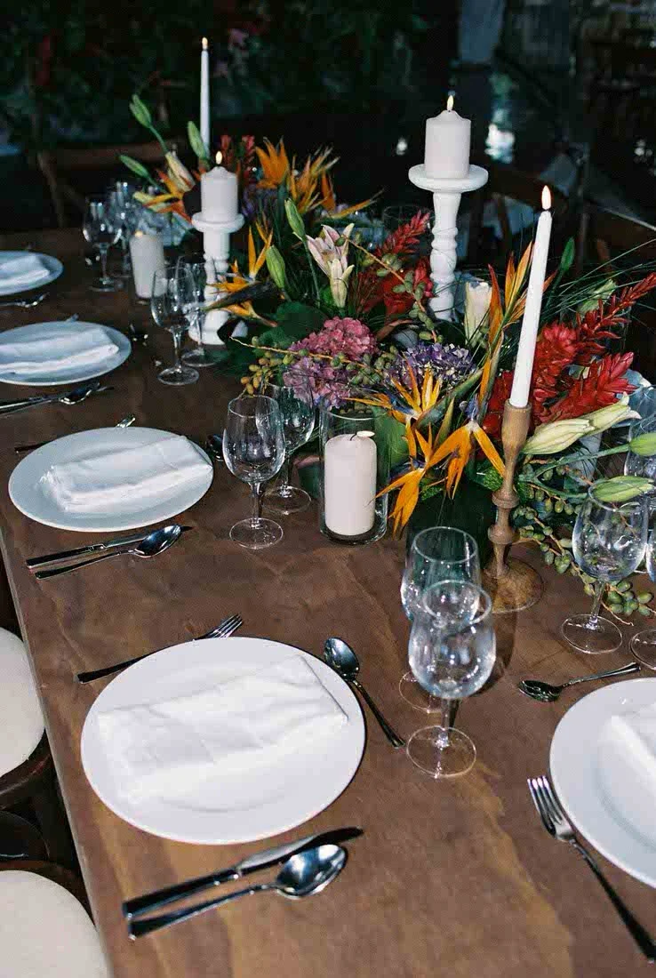 A long wooden dining table set for a formal event with white plates, silverware, wine glasses, and a floral centerpiece with candles.