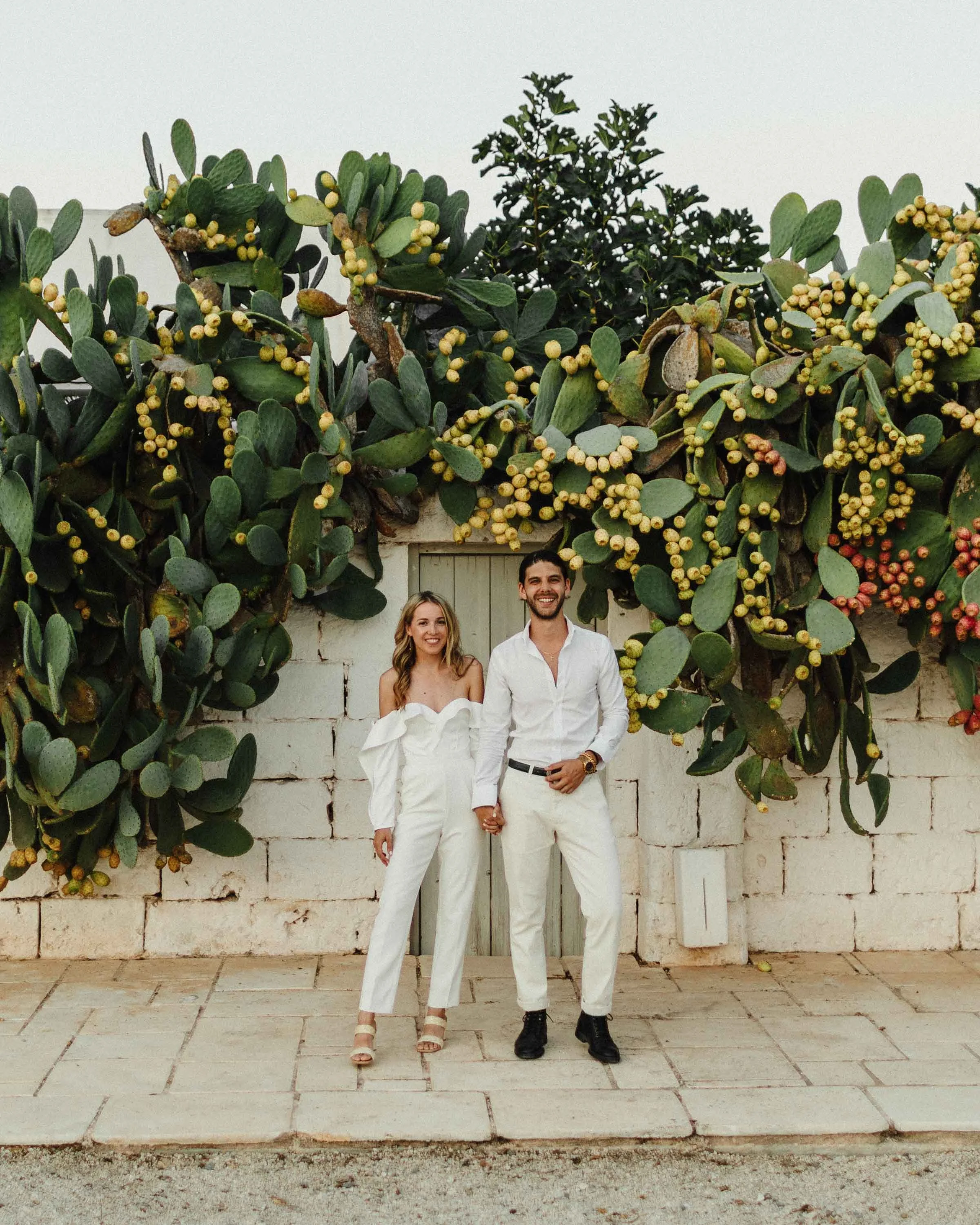A smiling couple holding hands in front of a wall with large cactus plants.