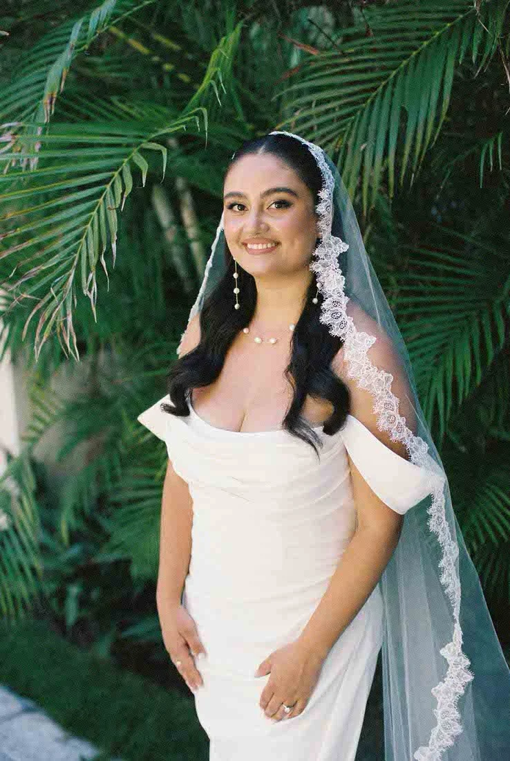 A bride in a white wedding dress with a lace-edged veil, smiling, standing outdoors near green palm leaves.
