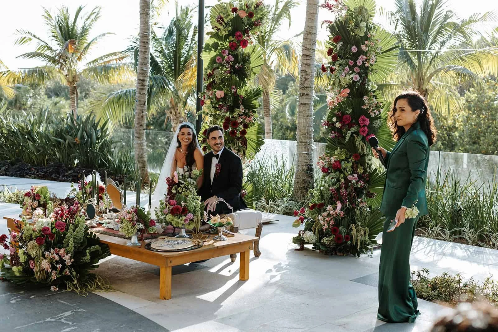 Bride and groom Laila and Arash seated at altar during wedding ceremony at Garza Blanca Cancun, officiant speaking, burgundy and blush floral installations with palm trees and ocean view in background