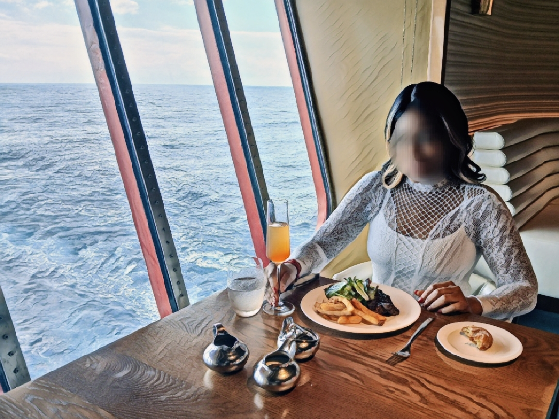 A woman with shoulder-length dark hair, sitting at a table on a cruise ship, enjoying a luxurious meal with ocean views through large windows.