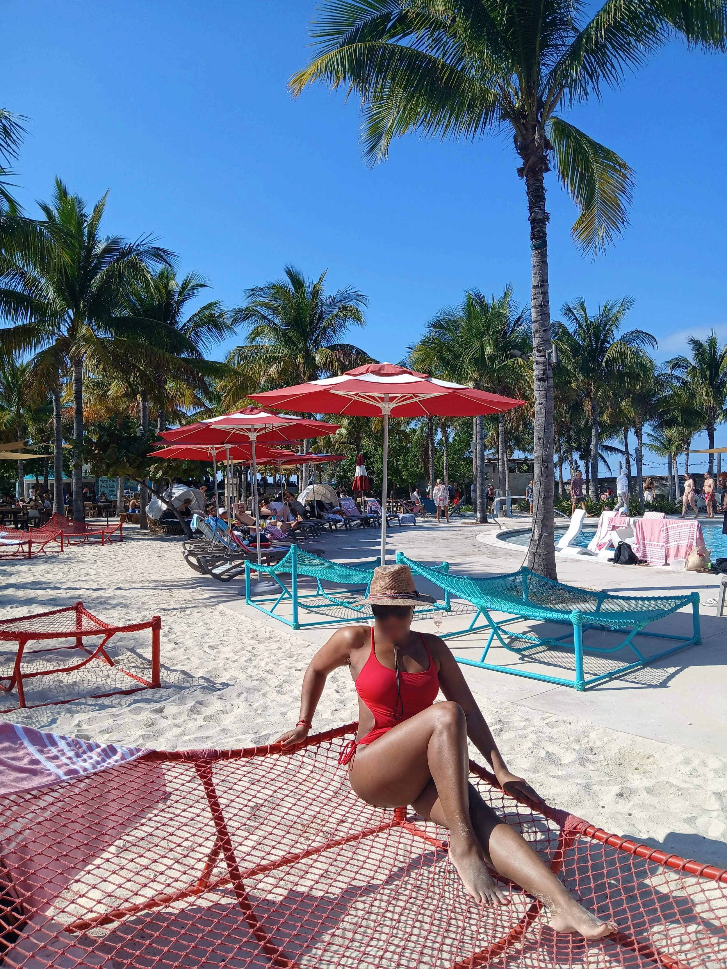 Sexy Woman in red swimsuit and wide-brimmed hat sitting on a netted lounge chair on a sandy beach with palm trees, umbrellas, and other beachgoers in the background.