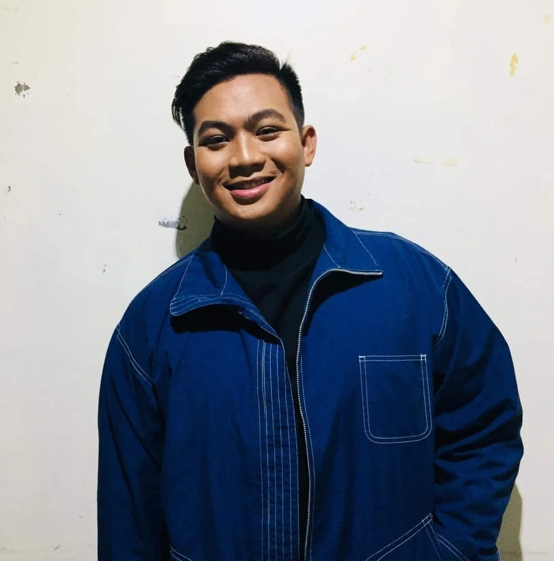 A young man smiling, wearing a black turtleneck and a blue jacket, standing against a plain off-white wall.