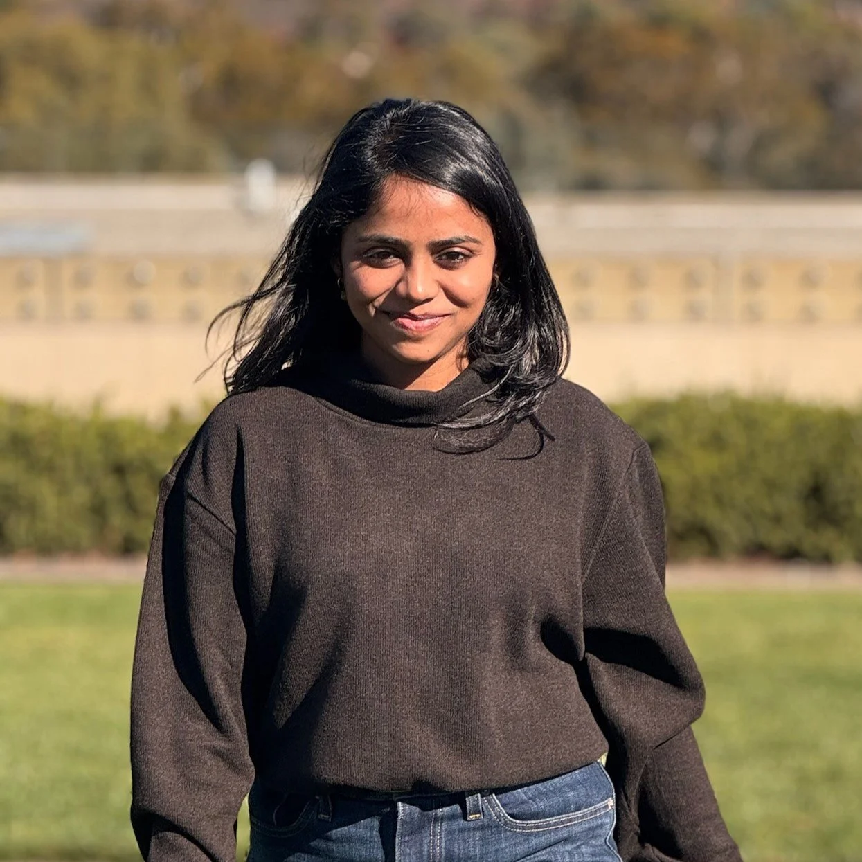 A woman with black hair and wearing a dark sweater standing outdoors in a park during daytime, smiling at the camera.