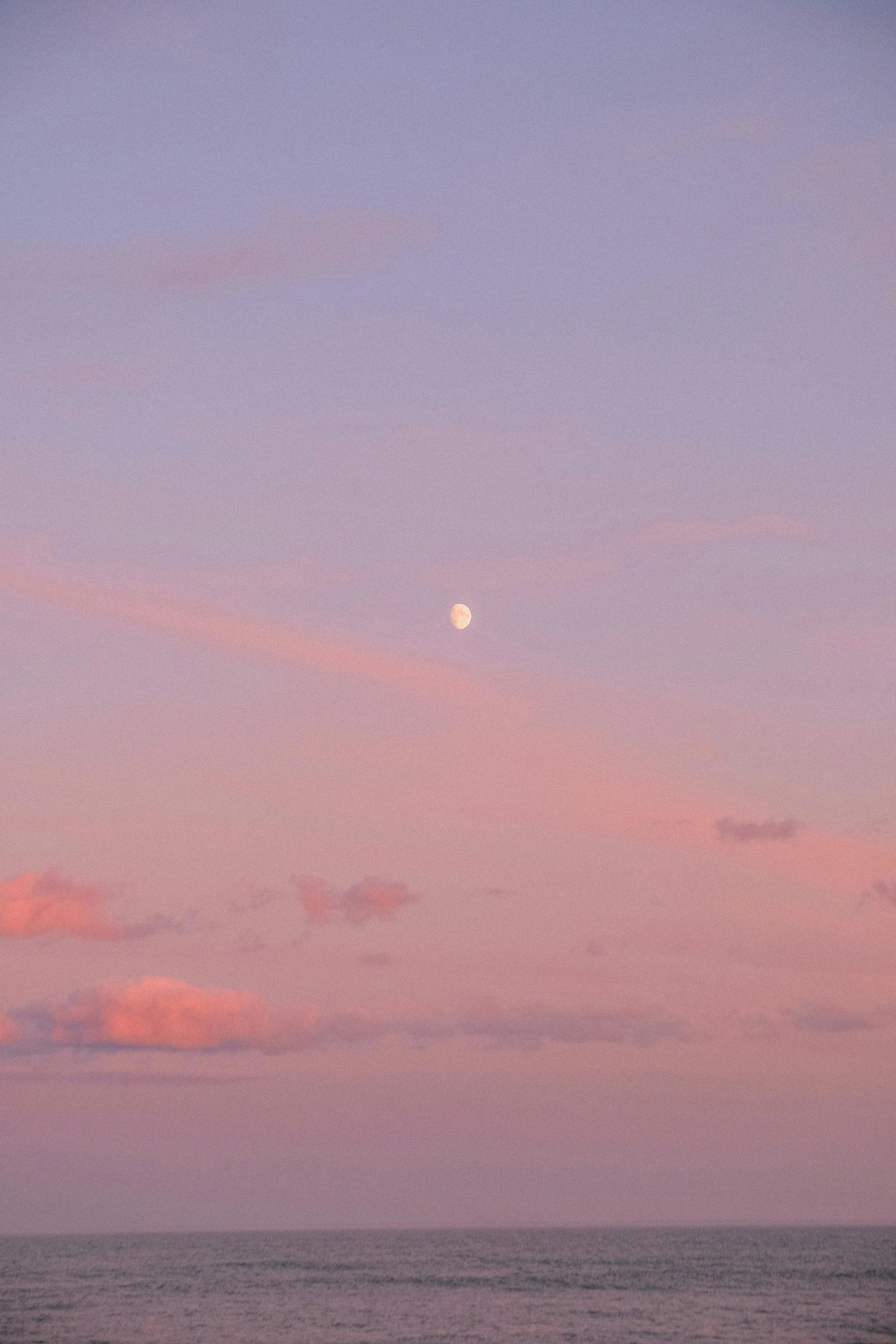 A peaceful ocean view during sunset with pink and purple clouds and the moon visible in the sky.