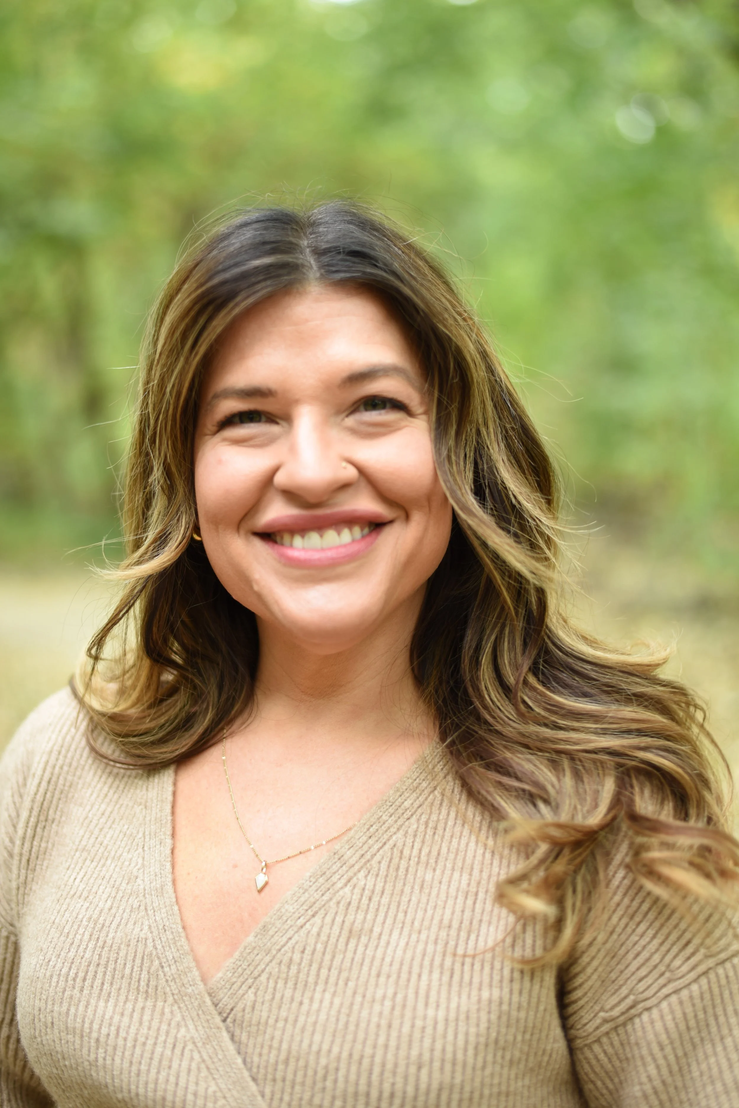 Therapist Holly Tofanelli with long wavy brown hair smiling outdoors with a blurred green forest background.