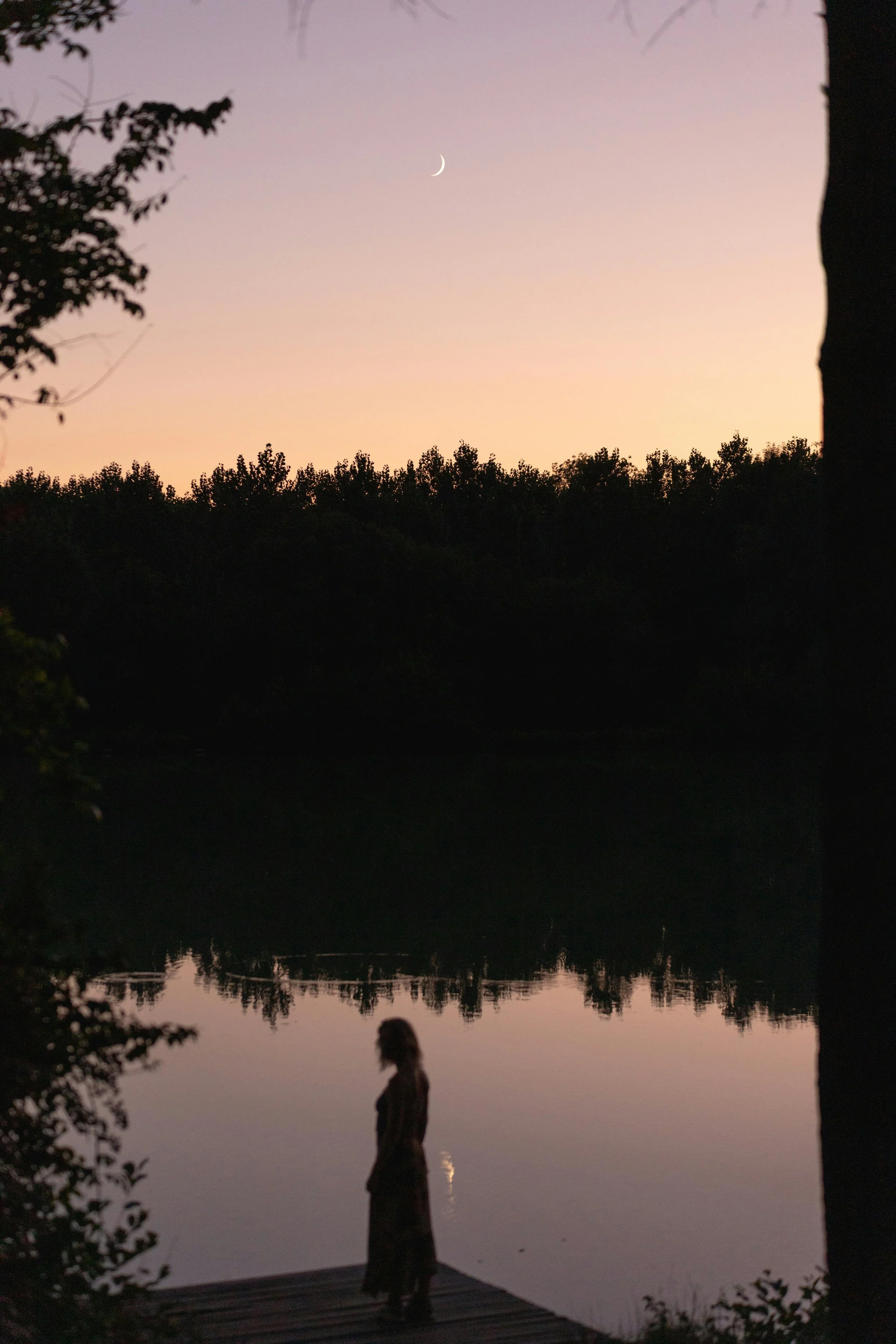 Silhouette of a person standing on a dock by a calm body of water during sunset, with a crescent moon visible in the sky and trees in the background.