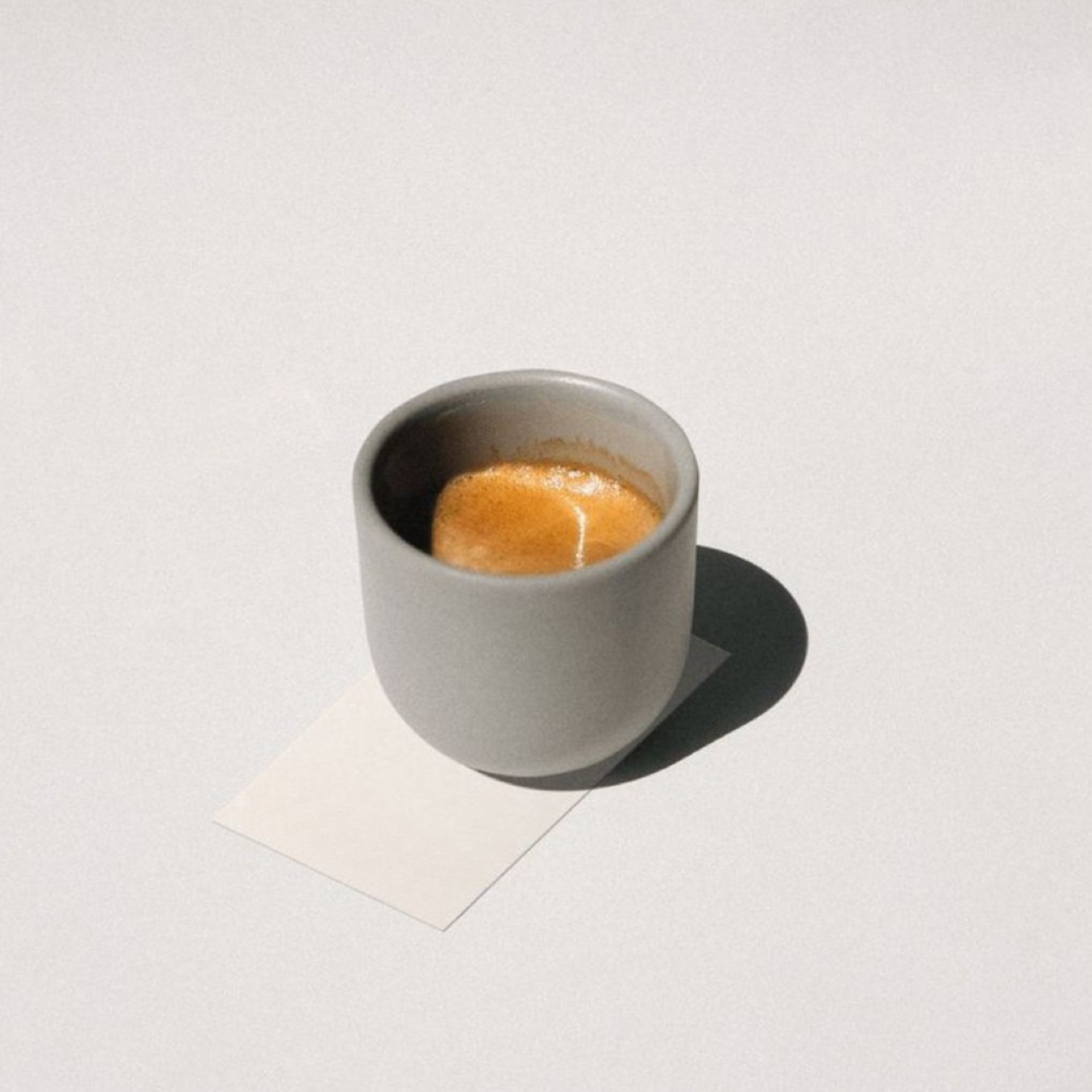A small white coffee cup filled with espresso, placed on a white surface with a white background. The cup casts a shadow on the surface.