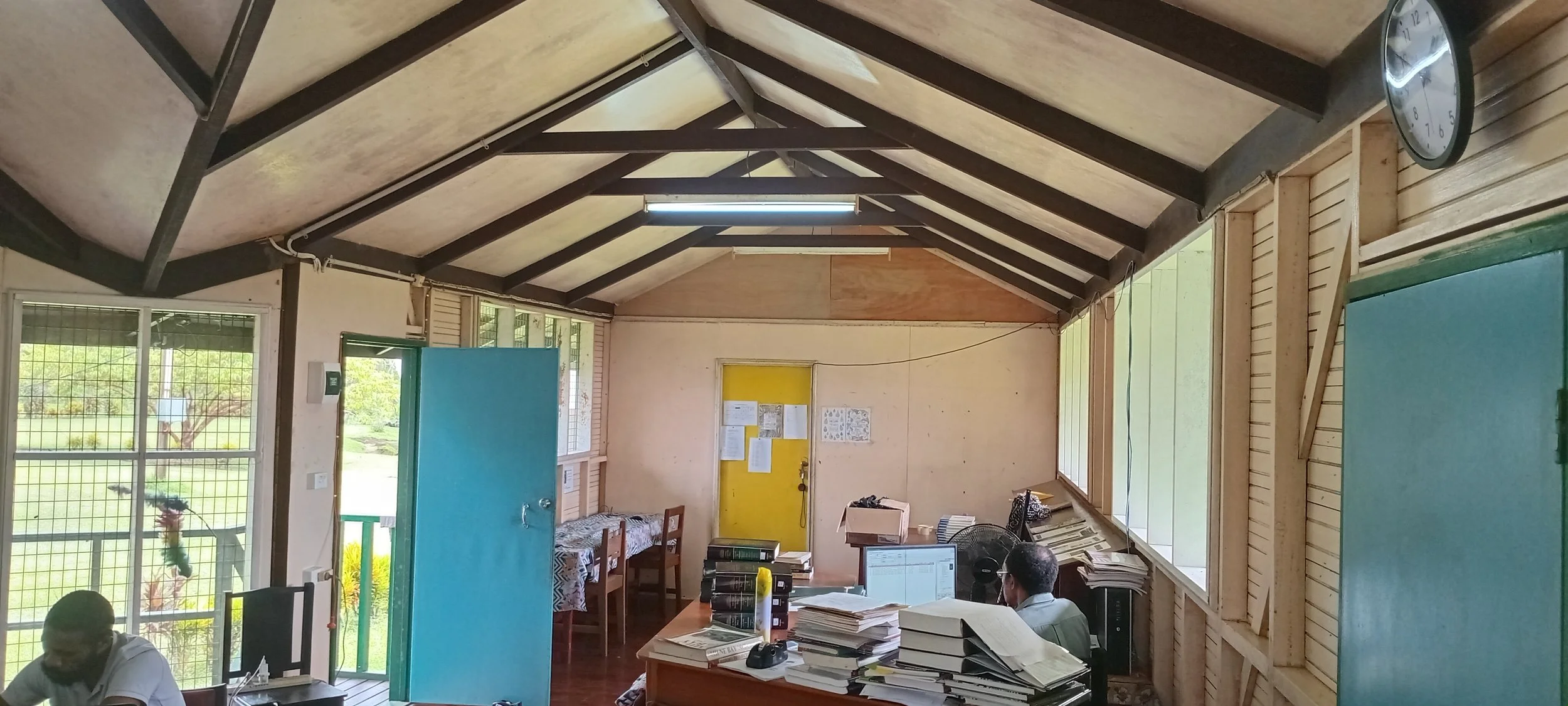 Interior of a small office or classroom with a yellow door, large windows, a table with stacks of papers, a person working at a computer, and clocks on the wall.