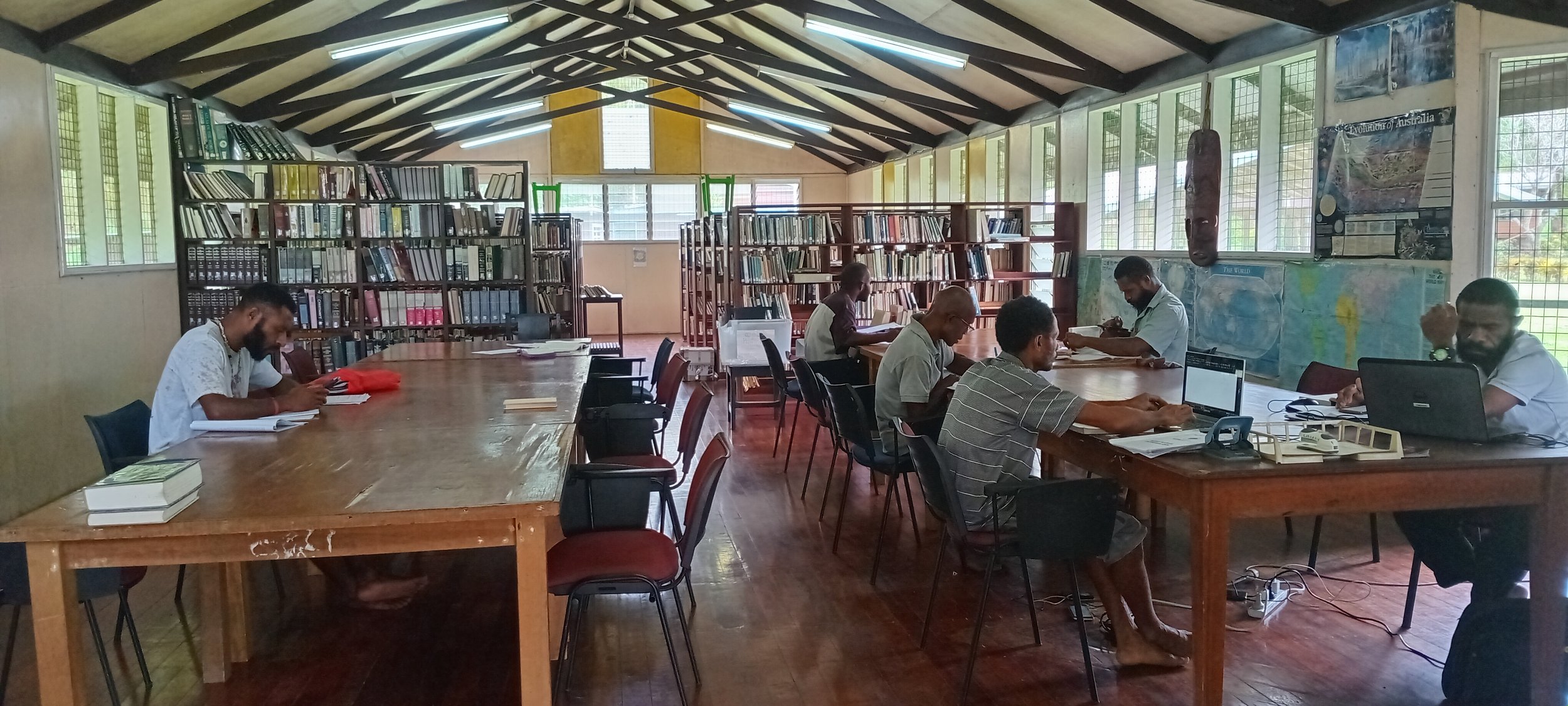 People sitting at tables studying or working in a library with bookshelves and maps on the walls.