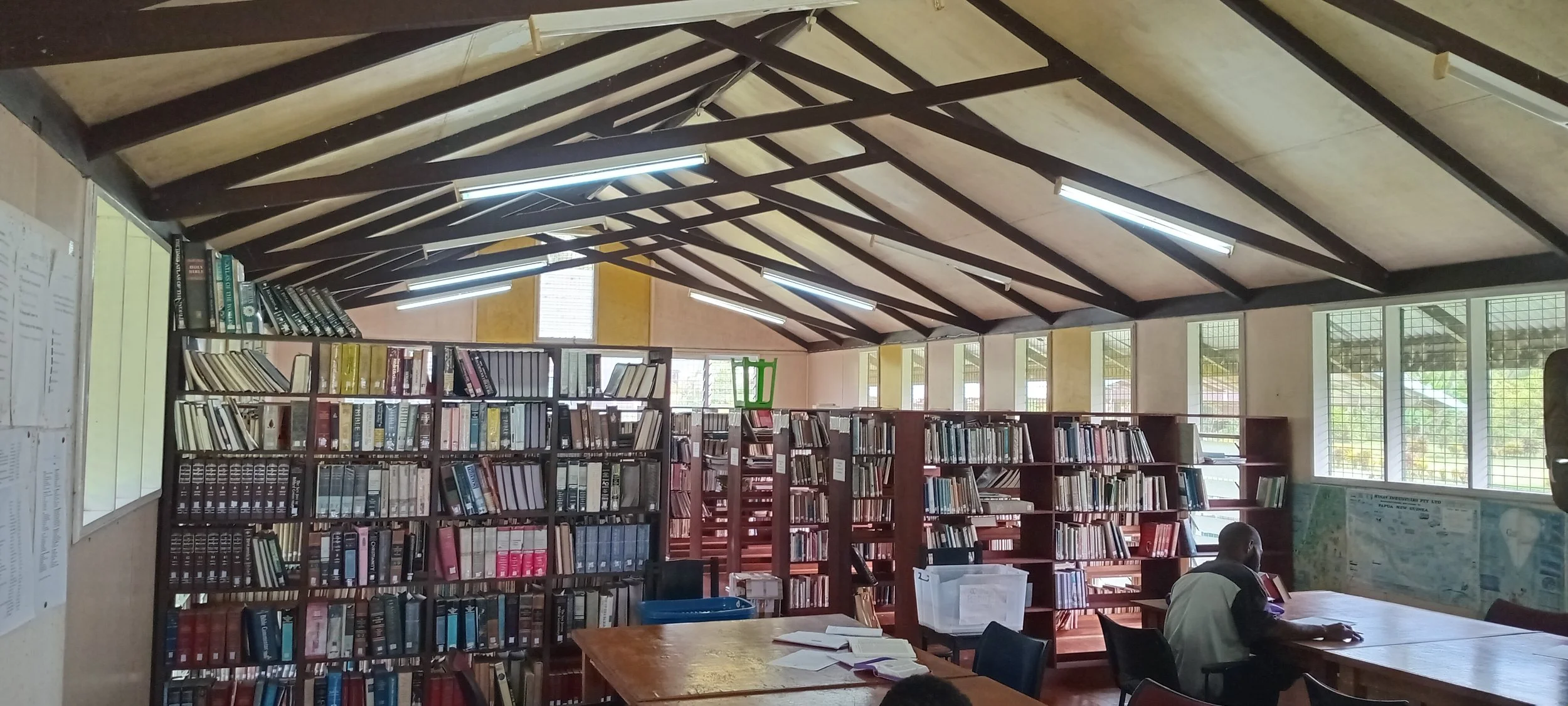 Inside a library with bookshelves, a man sitting at a table, and large windows letting in natural light.