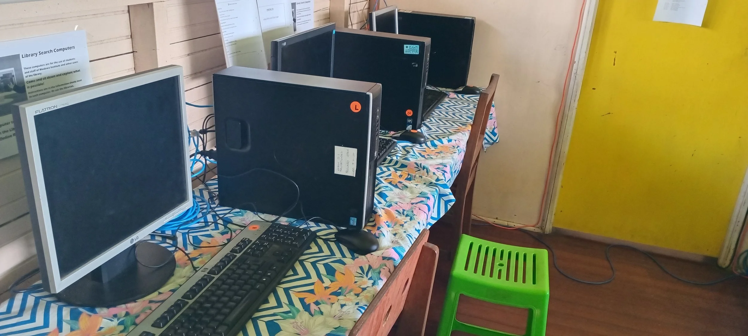 A row of four desktop computers on a colorful floral tablecloth on a wooden table, with a keyboard and mouse in front of the first computer, against a wall with notices and yellow door.
