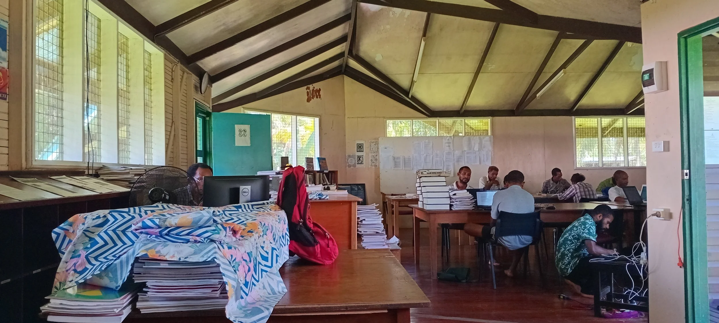 A classroom with desks, chairs, and people working on laptops and reading papers. There are large windows with mesh screens, a whiteboard, and stacks of papers and books on tables.