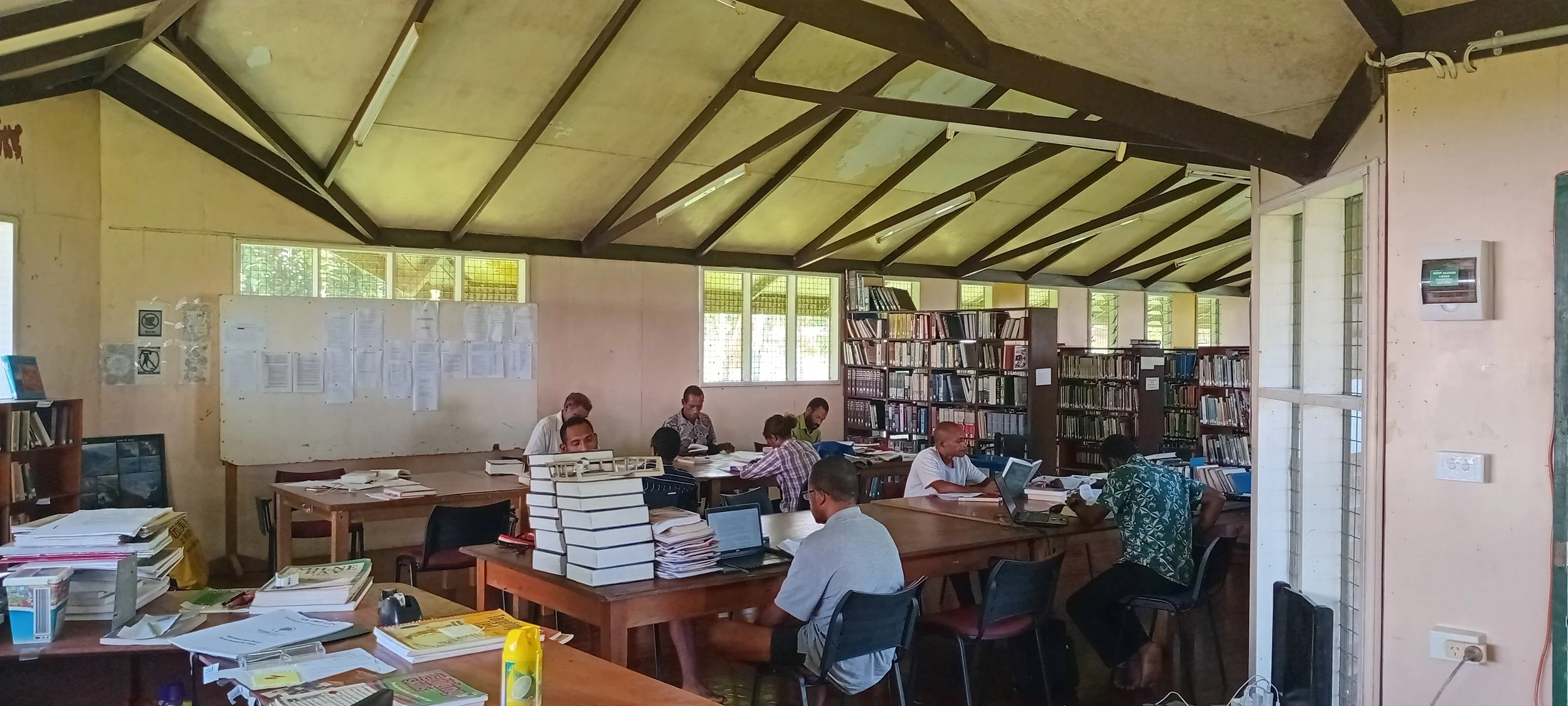 A crowded library room with multiple people working at tables, surrounded by bookshelves and papers.