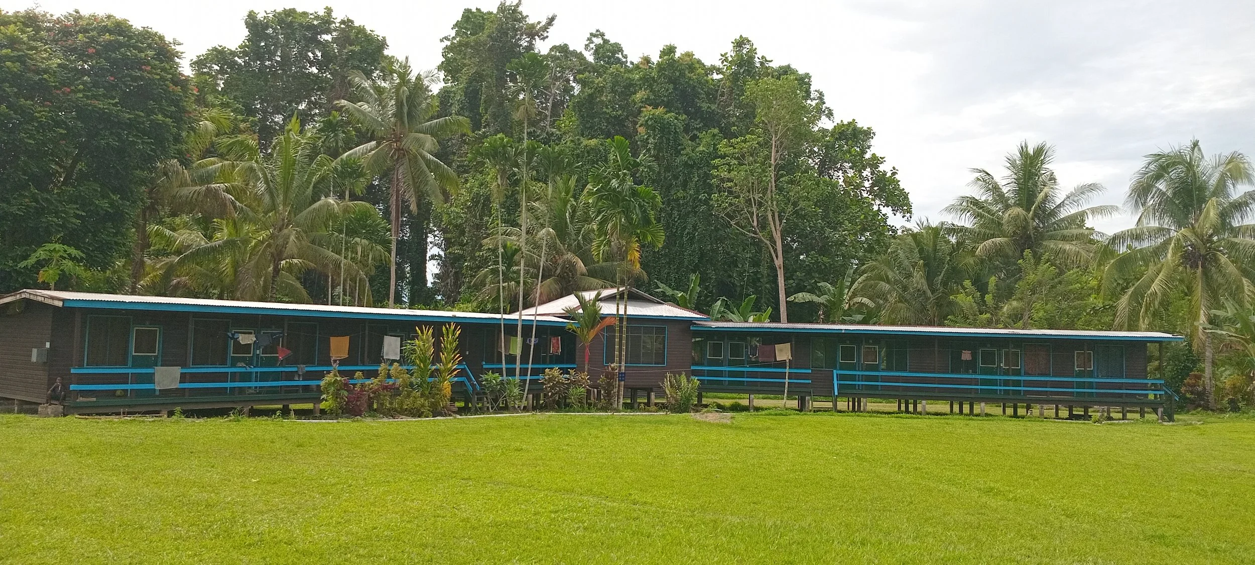 Single-story wooden building on stilts with a blue railing, surrounded by lush green grass and tropical palm trees, under a cloudy sky.