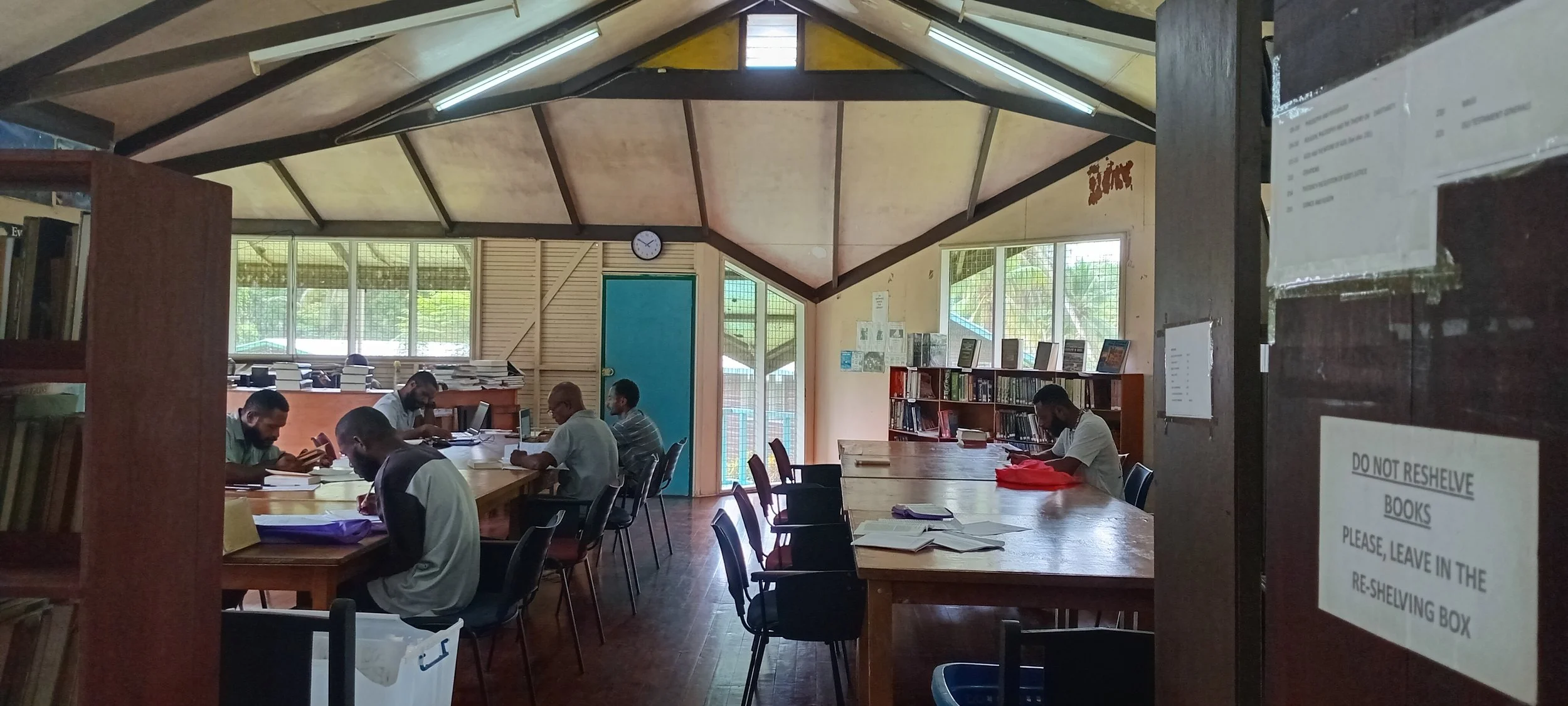 People sitting at tables reading and working in a library with wooden shelves and large windows.