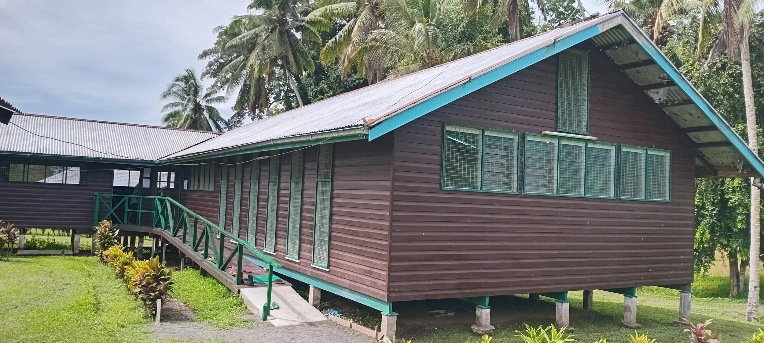 A wooden house on stilts with a metal roof, surrounded by palm trees and green foliage, with a small ramp leading to the front door.
