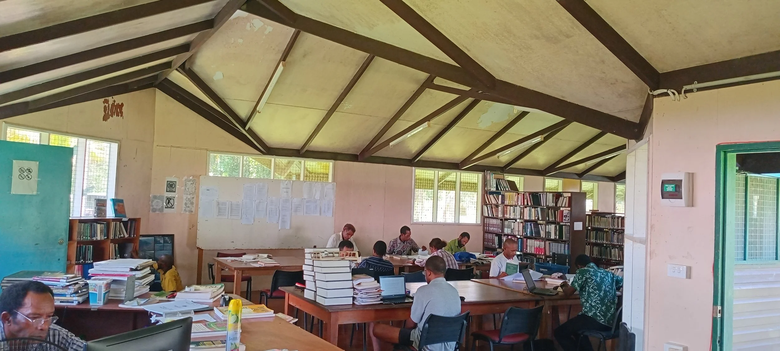 A library with several people studying at large tables, surrounded by bookshelves and stacks of books.