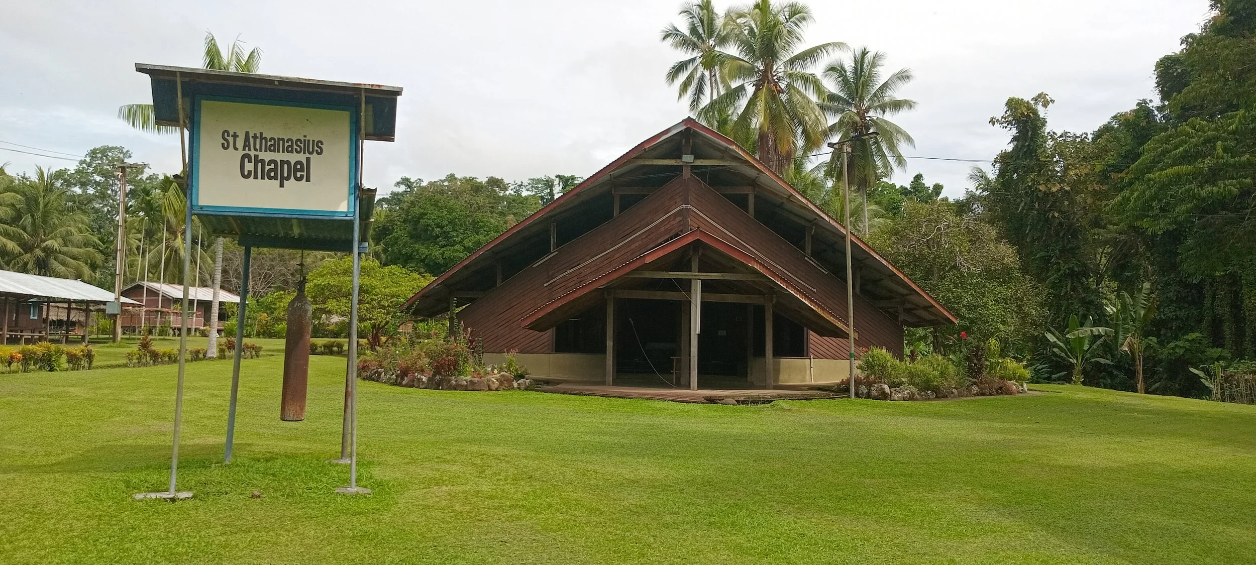 St. Athanasius Chapel, a wooden church-style building with a steeply sloped roof, surrounded by lush green grass, tropical trees, and shrubs. A sign indicating the chapel's name is visible in the foreground.