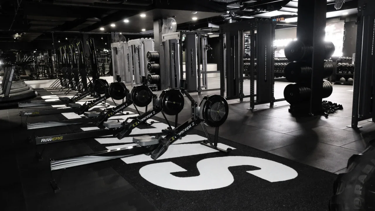 Empty gym with rowing machines in the foreground and various weightlifting equipment in the background, dimly lit with natural light