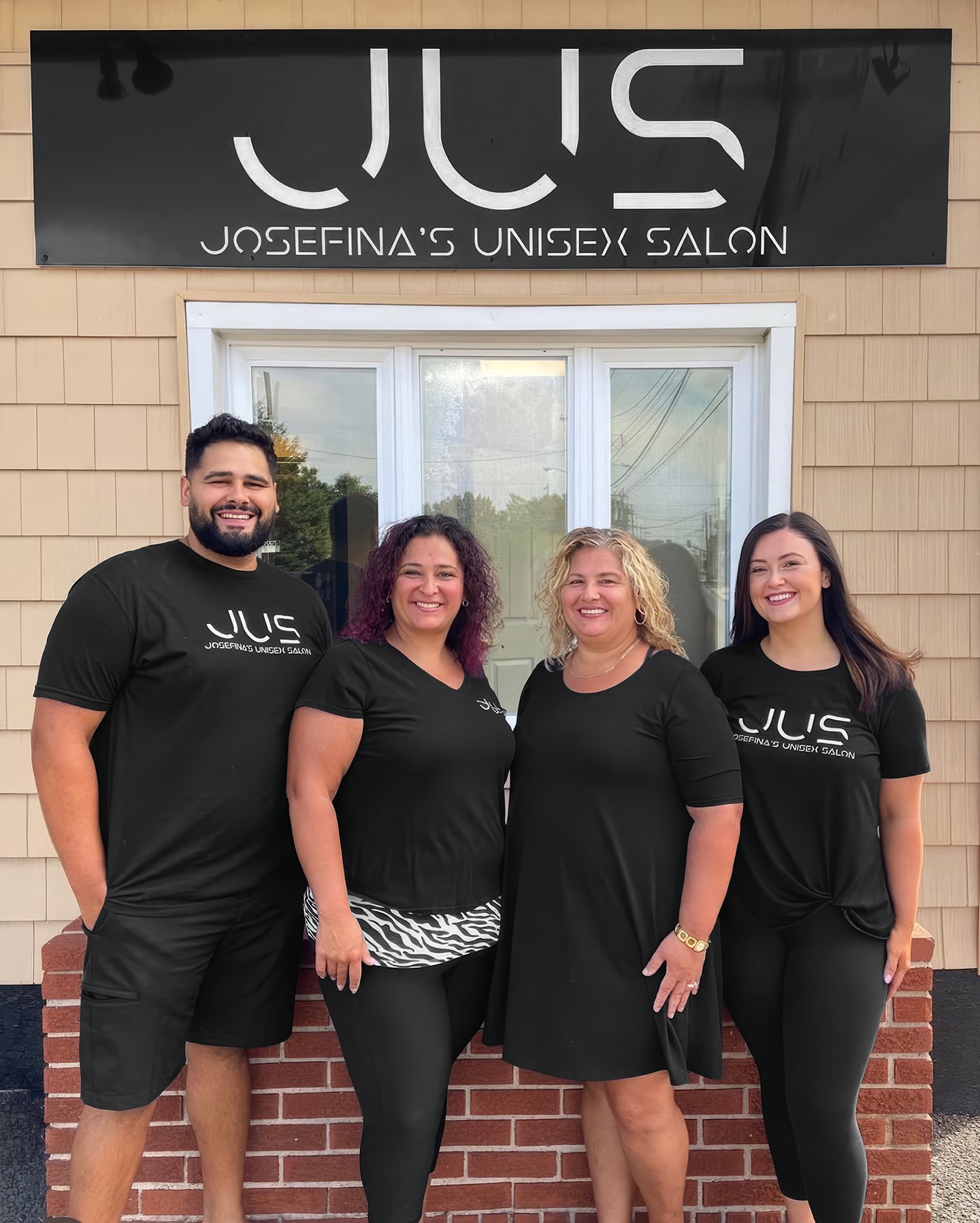 Three women and one man standing in front of a building with a sign that reads "JUS" and "Josefina's Unisex Salon," all wearing black shirts with the salon's logo. They are smiling and posing for the photo.
