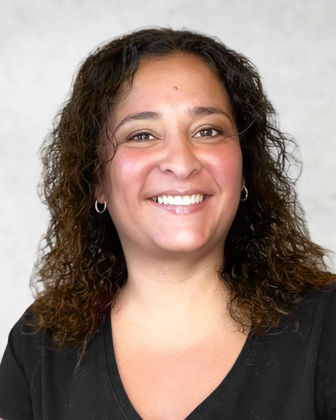 A woman with shoulder-length curly brown hair, smiling, wearing silver hoop earrings and a black top, against a plain light background.