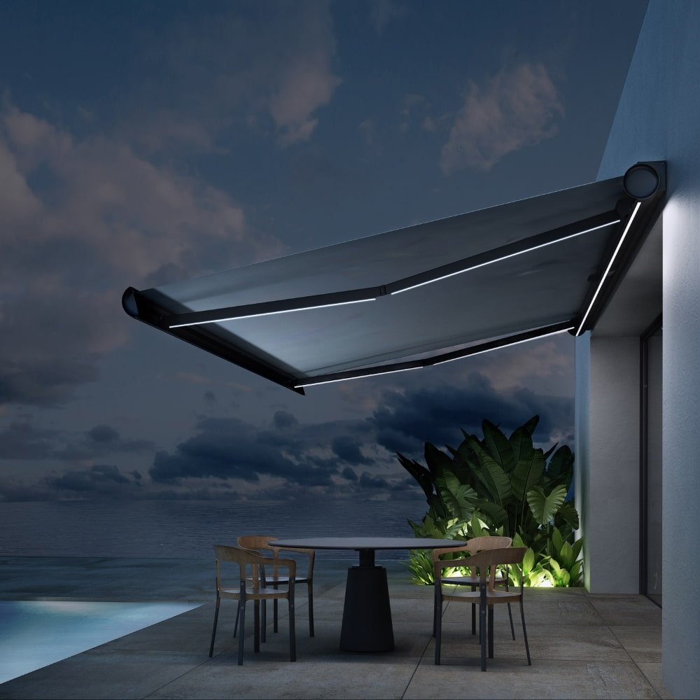 Exterior of a modern house with a folding arm awning extending from the side of the building. A round table with two chairs sits on a grey marble patio next to a pool and palm trees with a moody sunset sky