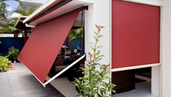 Exterior view of a white house with plants surrounding. Red extending arm awning