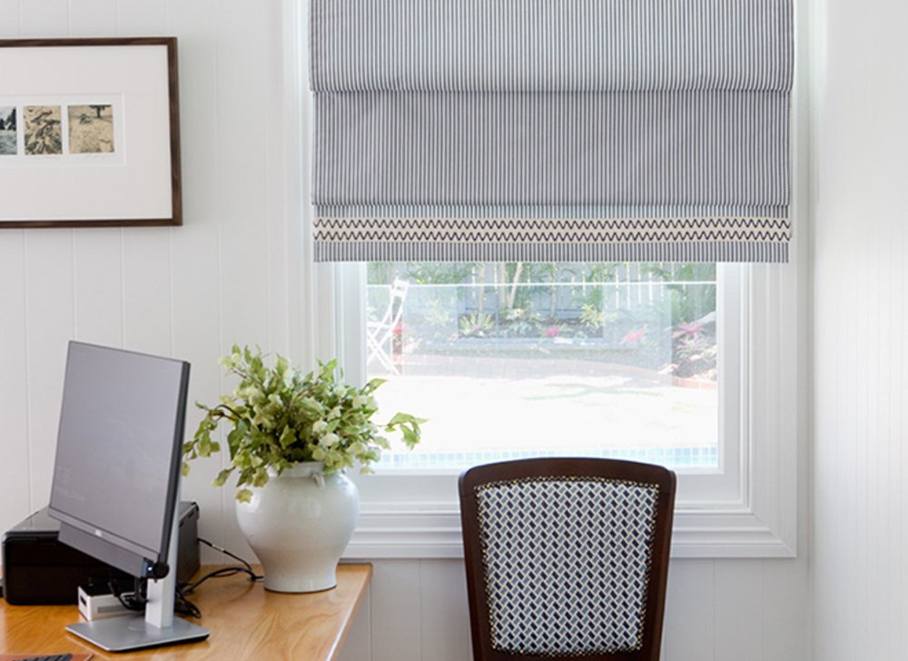 Grey and white striped roman blind in modern home office room. Computer on desk, white potted plant, photo frame, black desk chair, wooden desk, white window frame