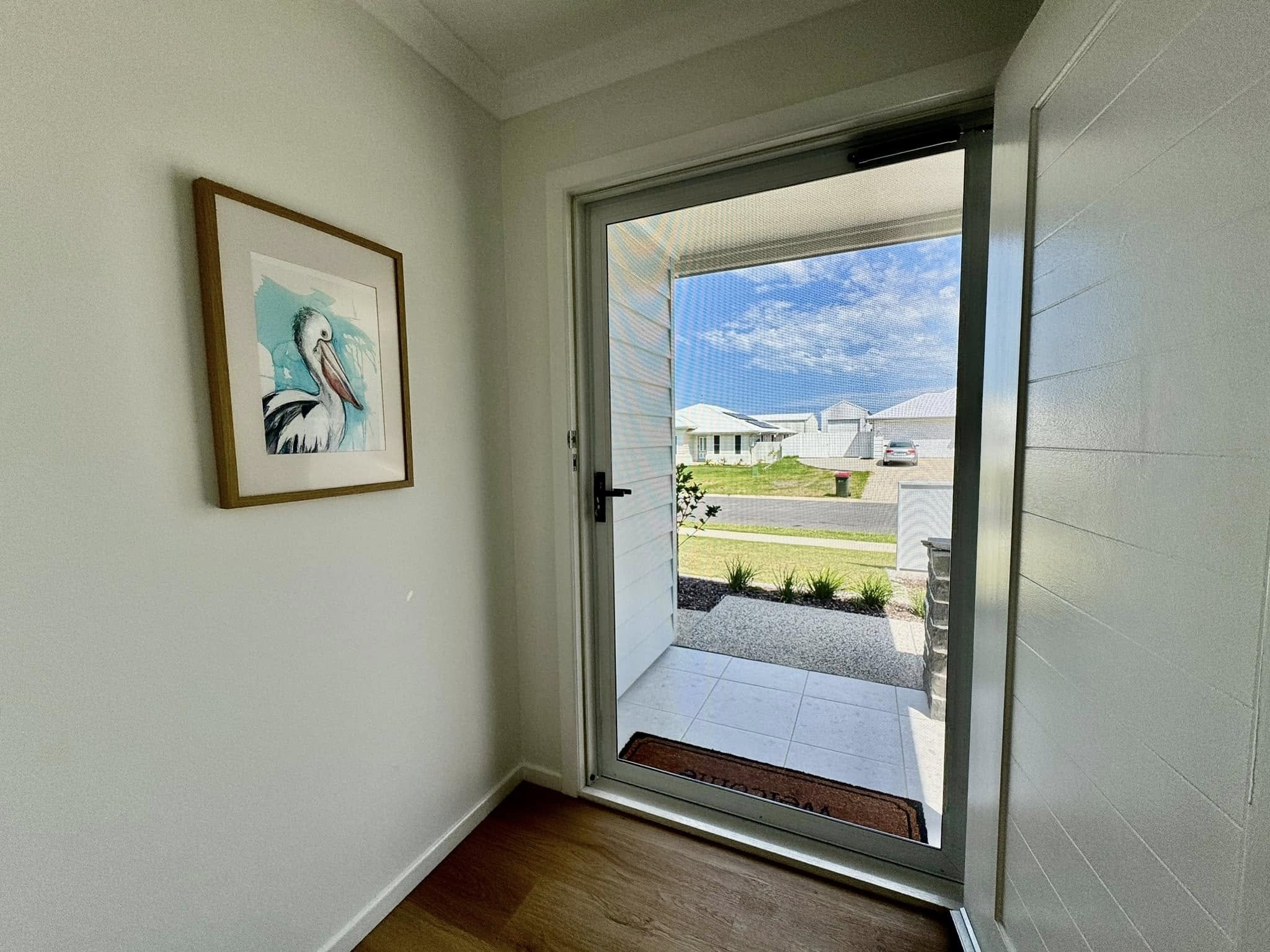 White hallway in a house with a flyscreen attached to the front door. Artwork of a pelican on the wall, view out to front garden with green grass and blue sky