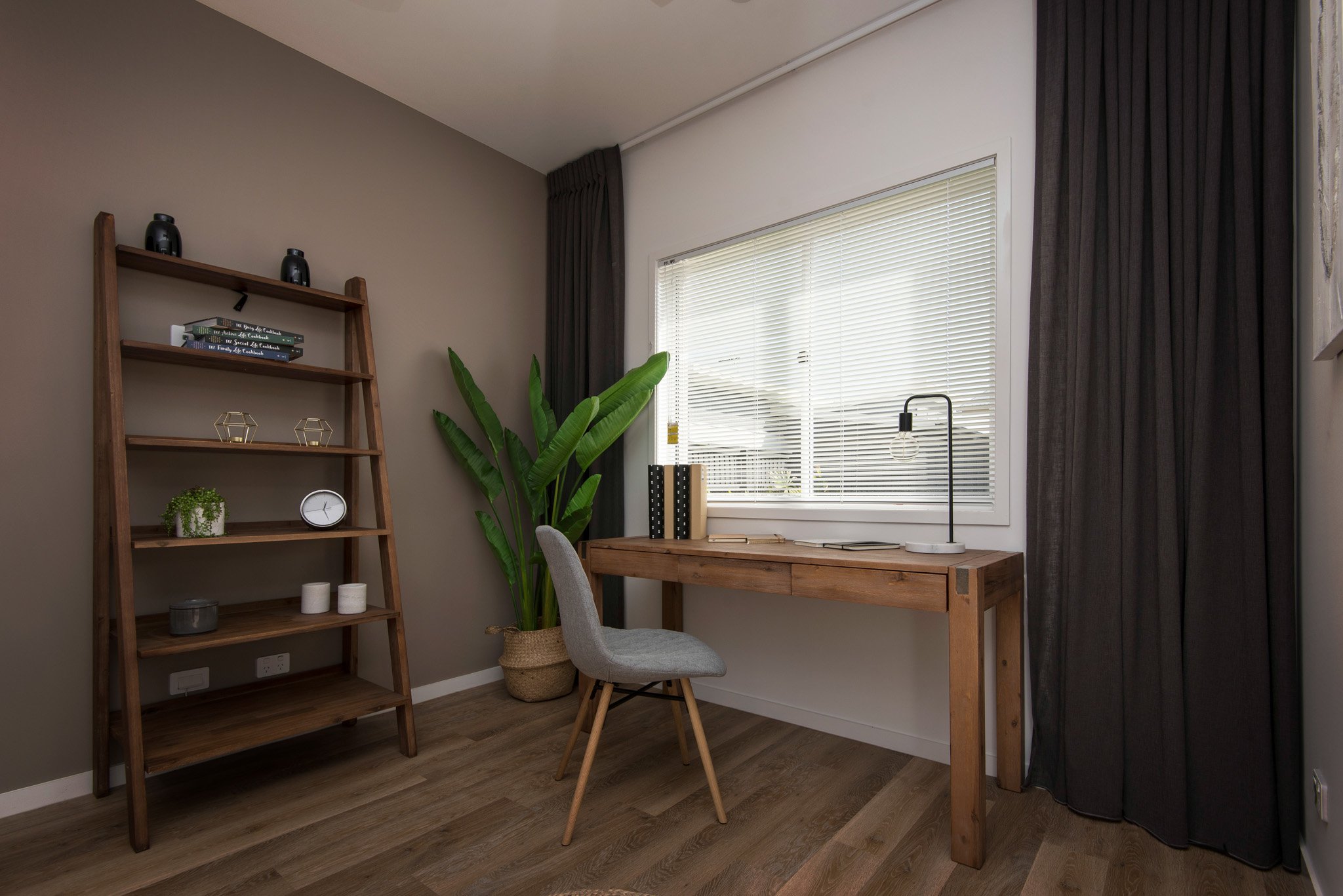 Venetian blind in a modern office space with taupe walls, wooden bookcase, wooden desk, grey chair, plant pot in corner, desk lamp and grey curtains
