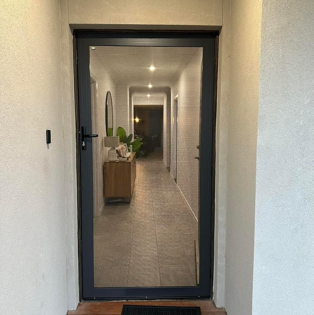 Black security screen attached to the front door of a house, looking down the corridor at white walls, potted plant and side unit