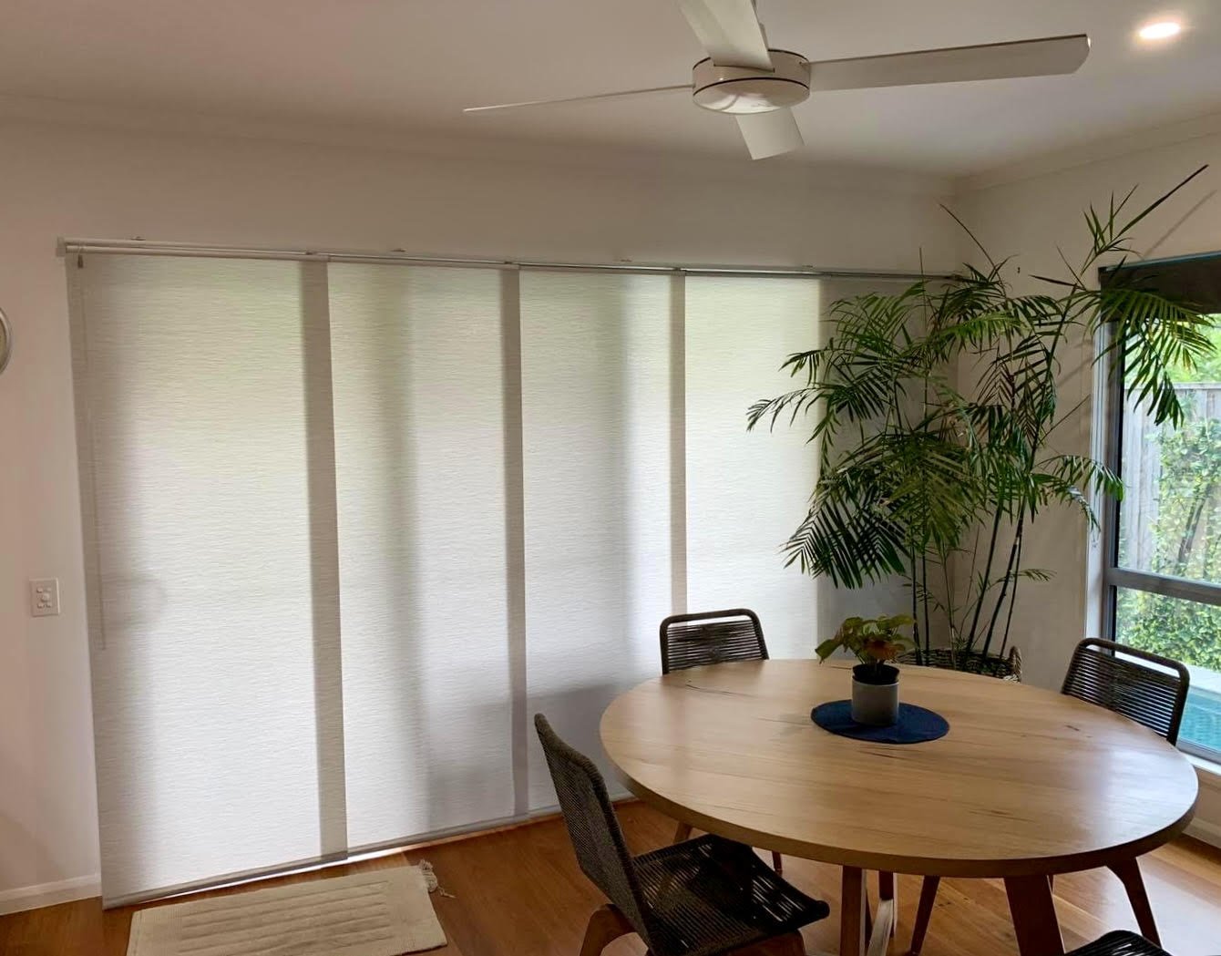 A dining room with a round wooden table, four chairs, a large potted plant near a window, and a white ceiling fan. The window has vertical blinds, and small rug is on the wooden floor. Installed panel glides by Sunburst, Central Queensland Australia