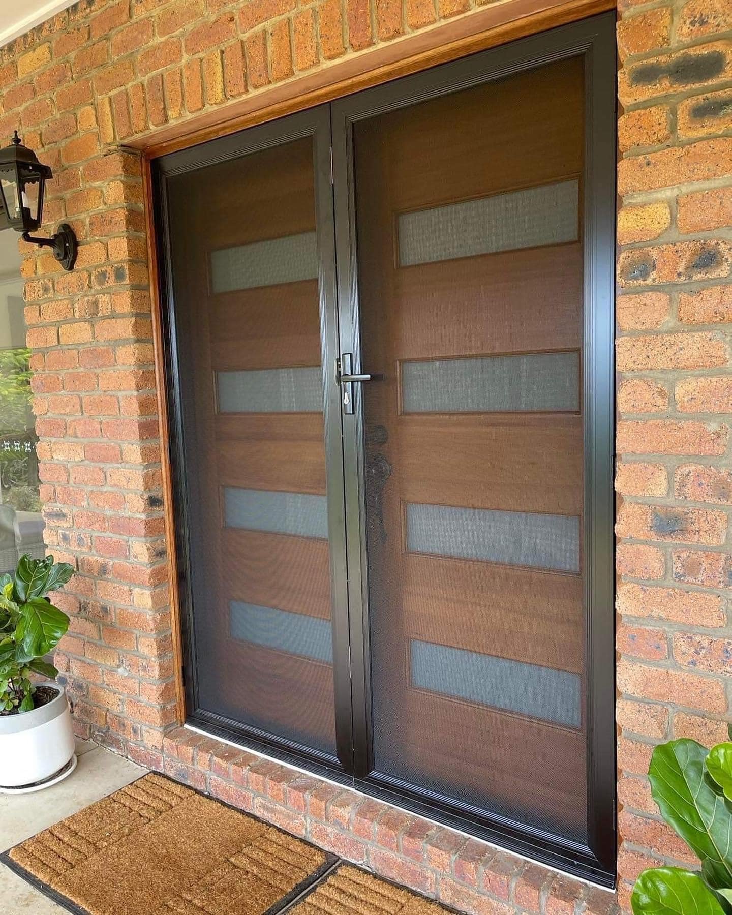 Brick house with door mat, potted fiddle leaf fig at either side of door, black wall sconce and black security screens over a wooden slatted door.