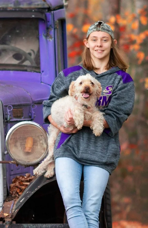 A girl with shoulder-length hair wearing a gray basketball cap backwards, a gray and purple hoodie, and light blue jeans, holding a cheerful cream-colored dog with a curly coat and open mouth, standing in front of a purple vintage truck with autumn leaves in the background.
